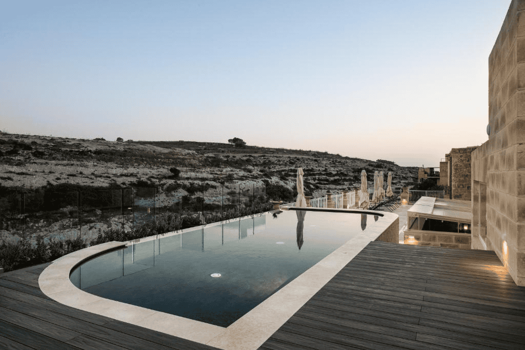 The terrace of Ta' Cesca Hotel, an infinity pool, and sun loungers look over a valley view.