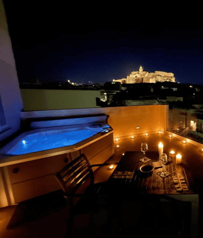 The candlelit terrace of the Cittadella View Penthouse, featuring a corner hot tub, dining area and a view of the floodlit Citadel.