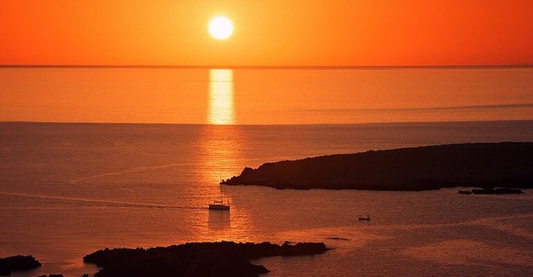 A tranquil sunset over the ocean with boats silhouetted against the vibrant horizon.